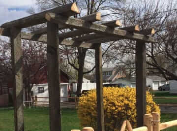 A wooden pergola stands in a grassy yard near a yellow flowering bush, with houses, trees, and a cloudy sky in the background. In front of the pergola, consider replacing the wooden fence with chain link fencing in Bergen County NJ.